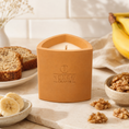 Load image into Gallery viewer, Candle in a terracotta holder with 'Texan Crafts' branding on a rustic background with bread, bananas, and nuts.
